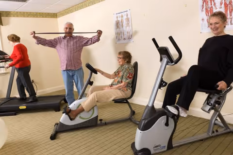 Four seniors exercising in a fitness room. One woman is walking on a treadmill, a man is stretching with a resistance band, and two women are using stationary exercise bikes. The room has beige walls with anatomical posters and a carpeted floor.