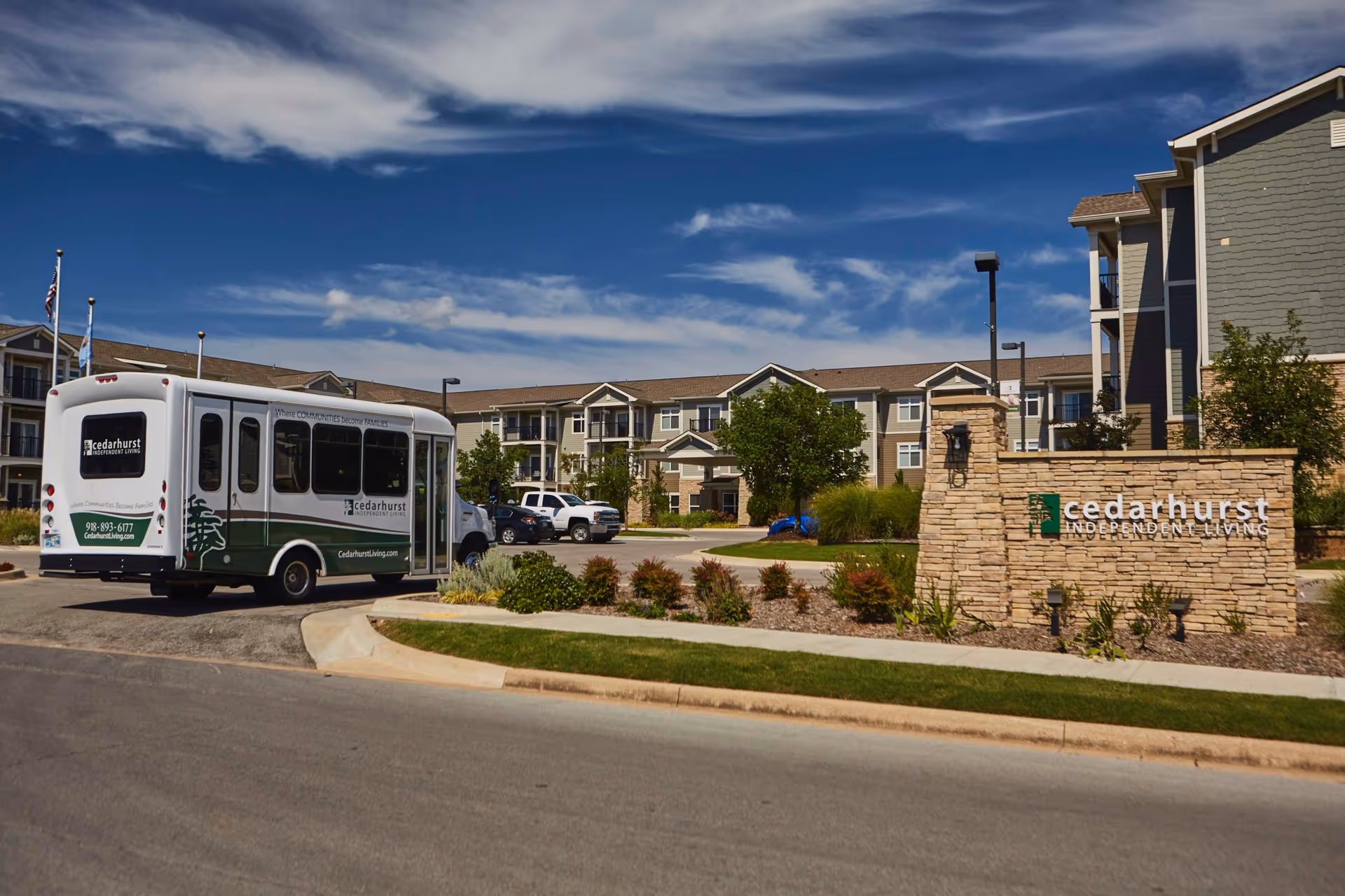 Exterior view of Cedarhurst Independent Living facility with a shuttle bus parked near the entrance. The building is a multi-story structure with balconies, surrounded by landscaped greenery and a clear blue sky with some clouds.