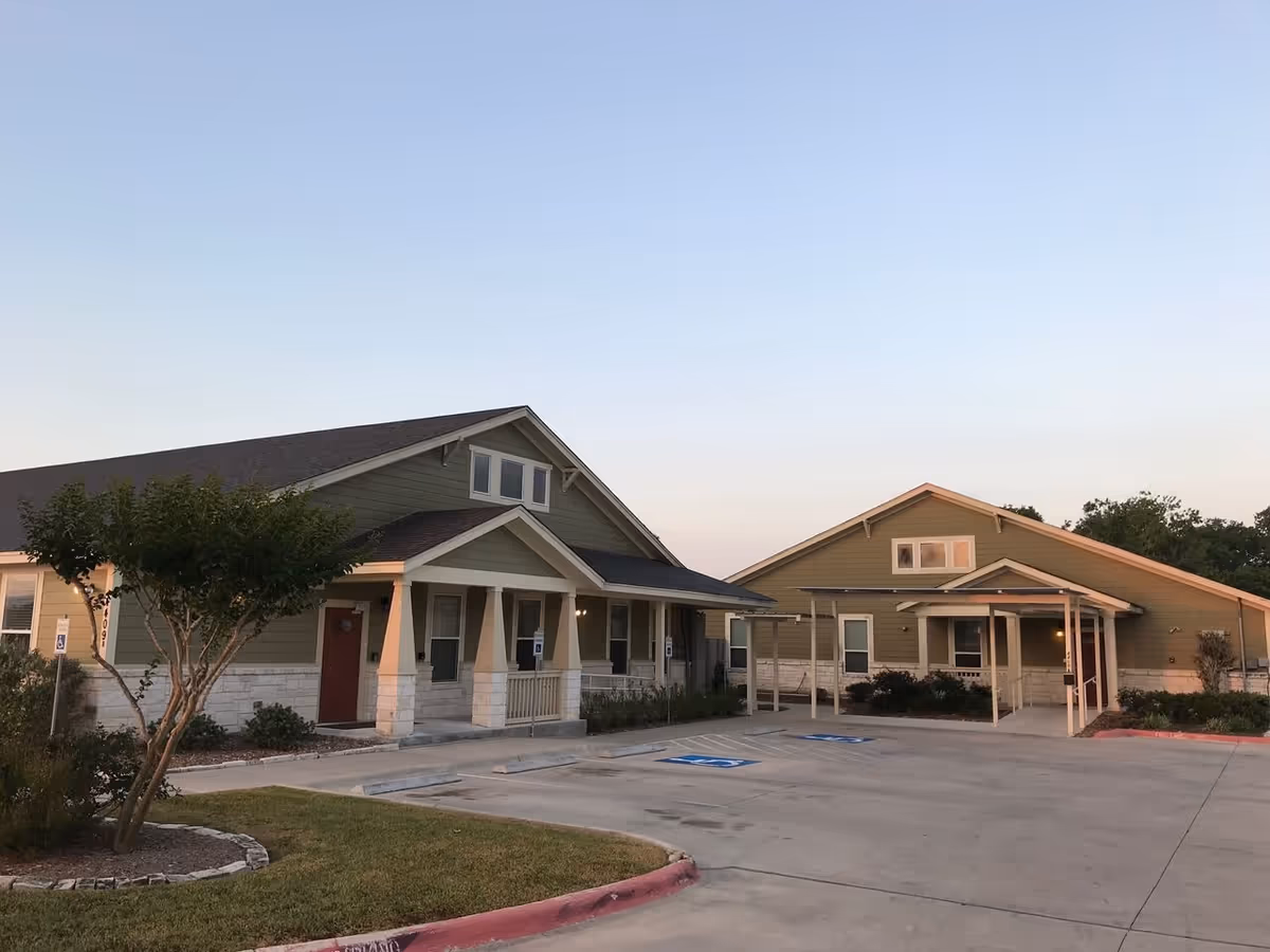Exterior view of Mercy House Victoria facility showing two single-story buildings with green siding and white stone bases, a small tree and landscaped area in front, and an empty parking lot with handicap parking spaces.