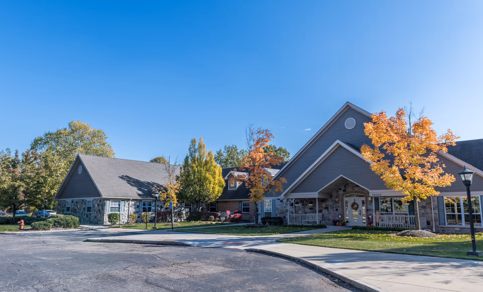 Exterior view of a senior living facility building with stone and siding facade, surrounded by trees with autumn foliage and a clear blue sky. There is a paved driveway and sidewalk in front of the building.