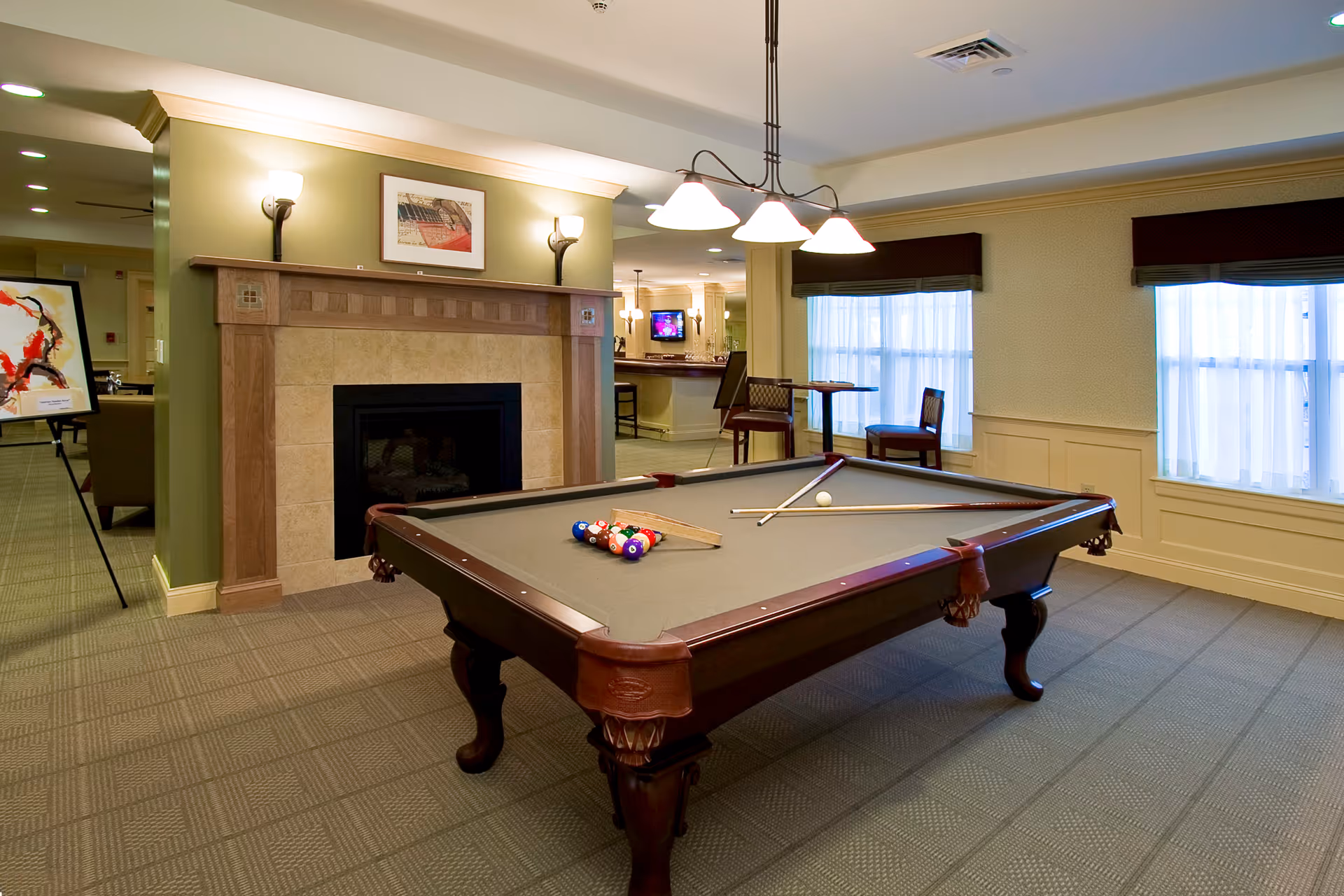 A well-lit room featuring a pool table with billiard balls and cues arranged on it. The room has a fireplace with a wooden mantle, two wall sconces, and a framed picture above the fireplace. There are two windows with sheer white curtains and dark valances. In the background, there is a small table with two chairs and a bar area with stools and a television mounted on the wall.