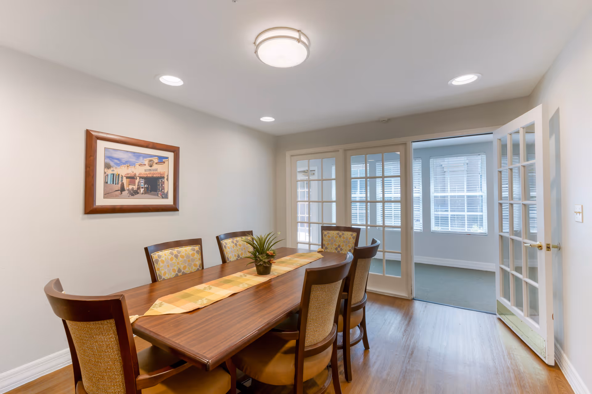 A dining room with a wooden table and six chairs, a decorative table runner, and a small plant centerpiece. The room has light-colored walls, a framed picture on the wall, recessed ceiling lights, and a ceiling-mounted light fixture. French doors with glass panes lead to another room with windows and blinds.