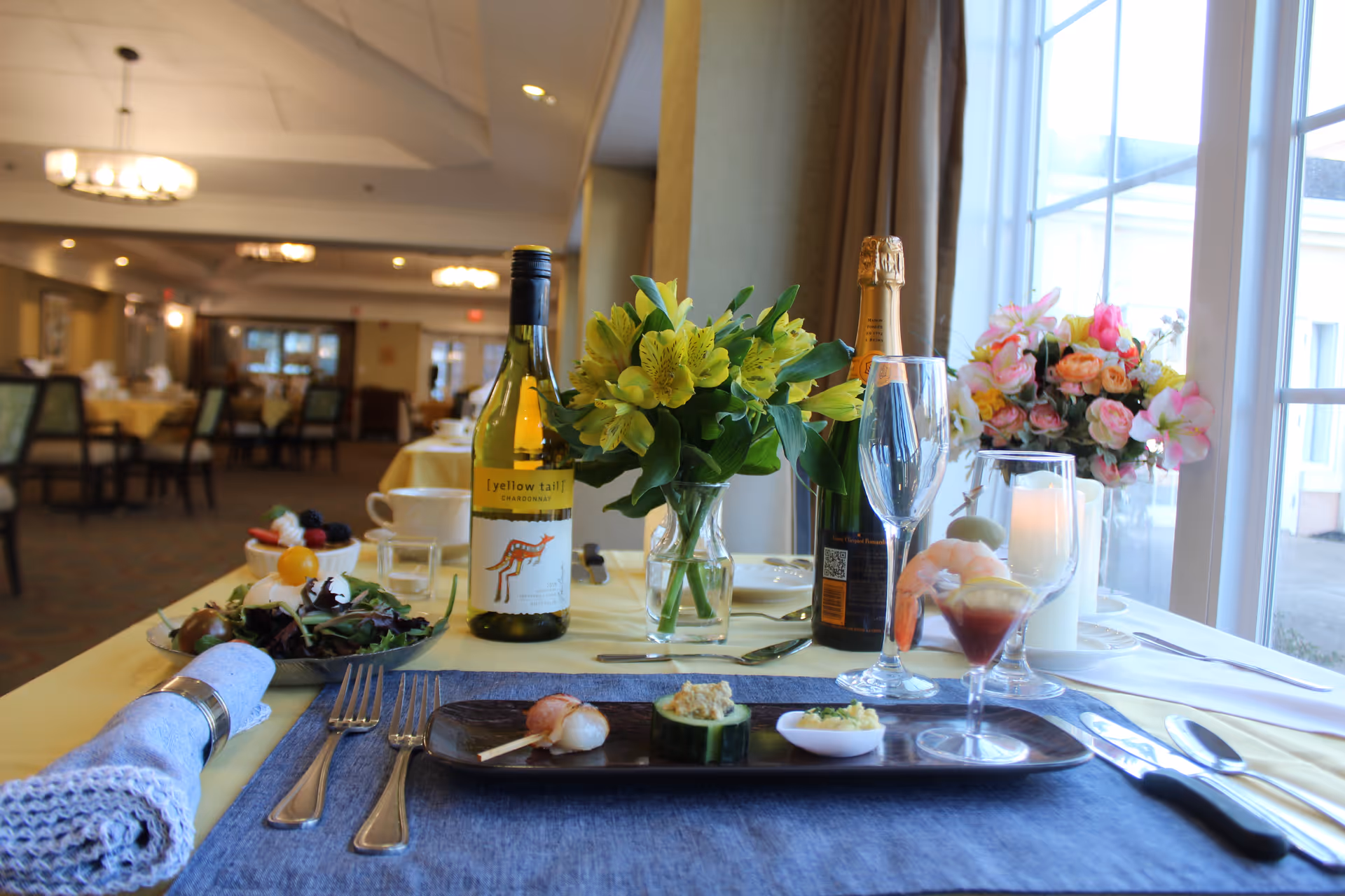 A dining table set with a blue placemat, silverware, a napkin with a ring, a plate with appetizers, a glass of wine, a bottle of Yellow Tail Chardonnay, a bottle of champagne, a vase with yellow flowers, and a vase with pink flowers near a large window in a dining room with multiple tables and chairs.