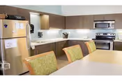 A modern kitchen with stainless steel appliances including a refrigerator, oven, and microwave. The kitchen has brown cabinets, a white countertop, and a dining table with green cushioned chairs in the foreground.
