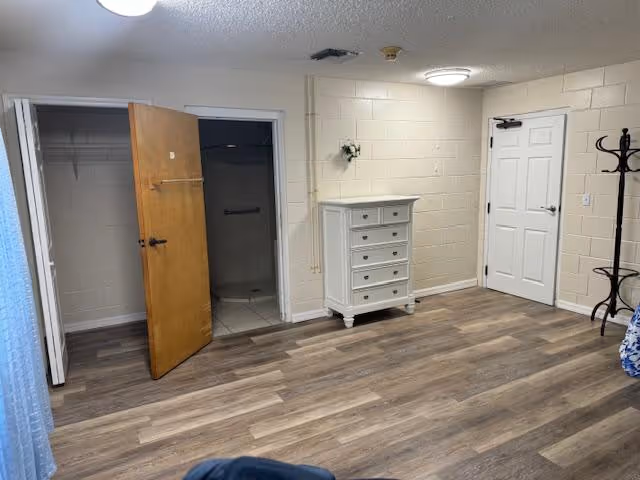 Interior room with wood-look flooring, beige painted cinder block walls, a white chest of drawers, an open closet with a wooden door, and a white door. A coat rack is visible on the right side near the door.