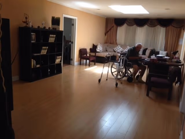 A spacious living room with wooden flooring, a black bookshelf filled with books and decorative items on the left, a beige sofa with patterned cushions near the window with curtains, and an elderly person sitting in a wheelchair at a table with chairs. A walker is positioned next to the wheelchair.