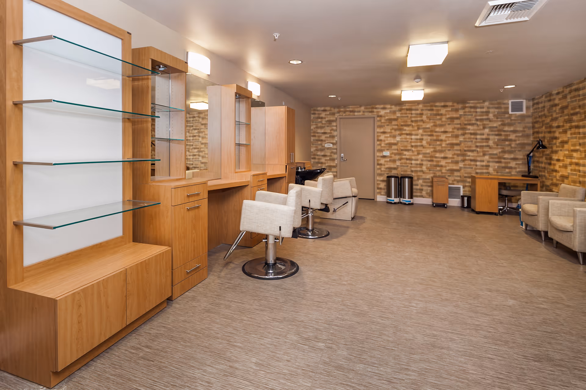 Interior view of a salon area in a senior living facility with wooden cabinetry, glass shelves, mirrors, salon chairs, and a washing station. The room has a textured brick-patterned wall, beige flooring, and additional seating with armchairs and a desk in the background.