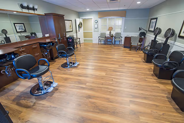 Interior view of a salon area in a senior living facility with wooden flooring, several black salon chairs, hair drying stations, and a counter with mirrors and lighting. There are also chairs and a small table near a window with blinds in the background.