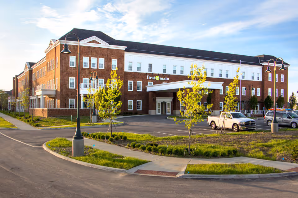 Three-story brick senior living building with a covered main entrance, parking lot, and young trees under a blue sky.