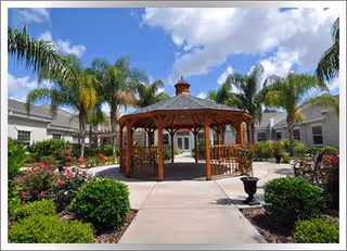 Outdoor courtyard with a wooden gazebo, palm trees, and manicured flowerbeds around concrete walkways.