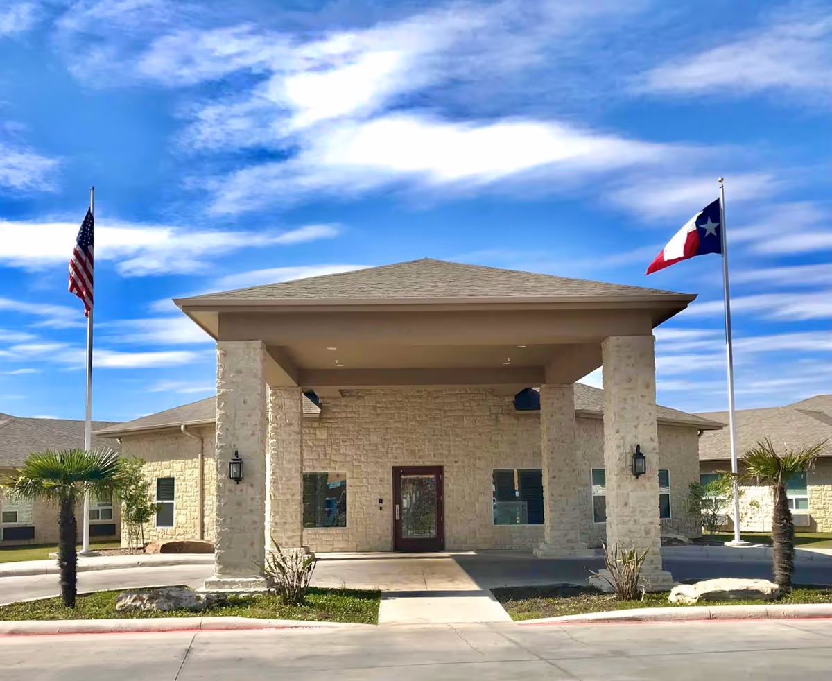 Front entrance of a stone-faced senior living facility with a covered portico and two flagpoles (American and Texas) under a blue sky.