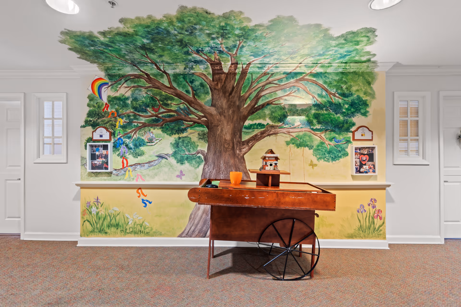 An indoor hallway area featuring a large mural of a tree with green leaves and a brown trunk painted on the wall. The mural includes colorful elements like a rainbow kite, butterflies, and flowers. In front of the mural is a wooden cart with a small model house and an orange cup on top. The walls are white with two small windows on either side of the mural.