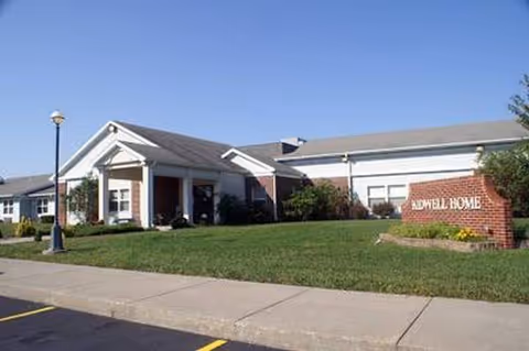Exterior view of a single-story senior care facility with a brick sign and lawn.