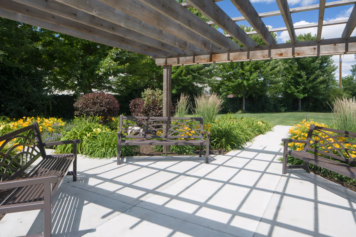 Outdoor seating area with three metal benches under a wooden pergola casting shadows on the concrete ground, surrounded by green bushes, flowering plants, and trees under a blue sky with some clouds.