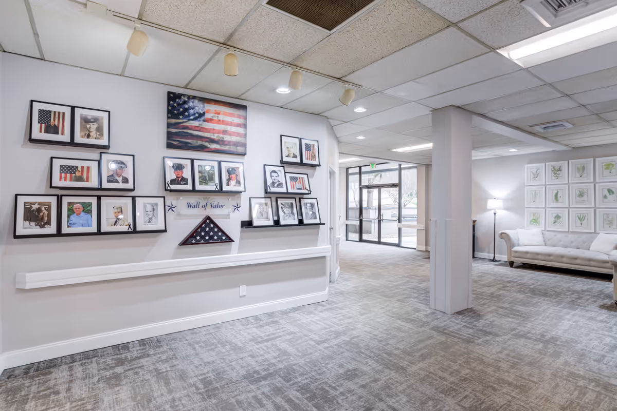 Bright senior living facility lobby featuring a 'Wall of Valor' photo display on the left and a seating area with a sofa and lamp on the right.
