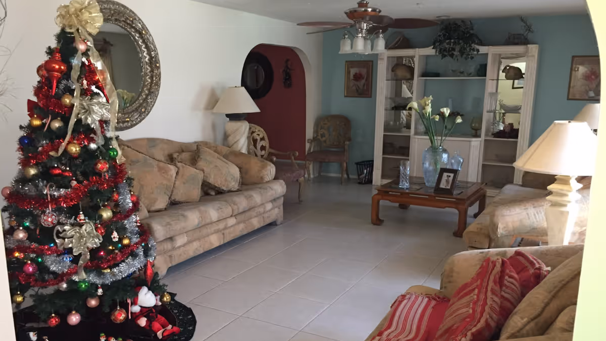 A cozy living room decorated for Christmas with a decorated Christmas tree on the left side. The room features beige sofas with cushions, a wooden coffee table with a vase of flowers, and a white shelving unit with decorative items. There are two table lamps, a round mirror on the wall, and framed artwork. The floor is tiled and the walls are painted in light colors with an archway leading to another room.