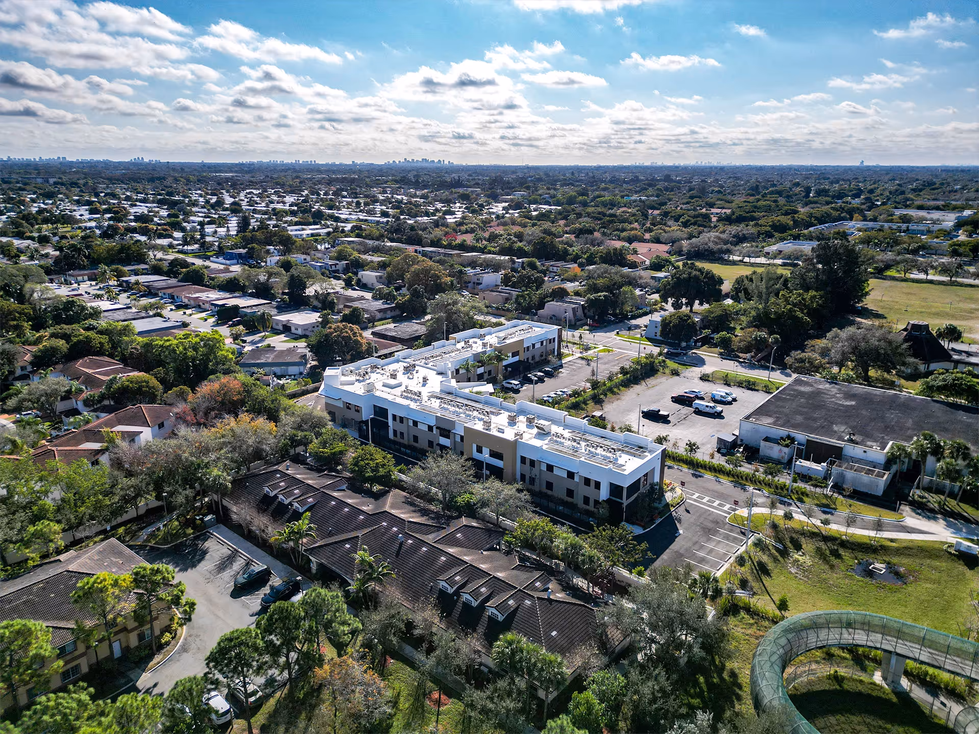 Aerial view of a low-rise senior living building surrounded by residential homes, parking areas, and trees under a partly cloudy sky.