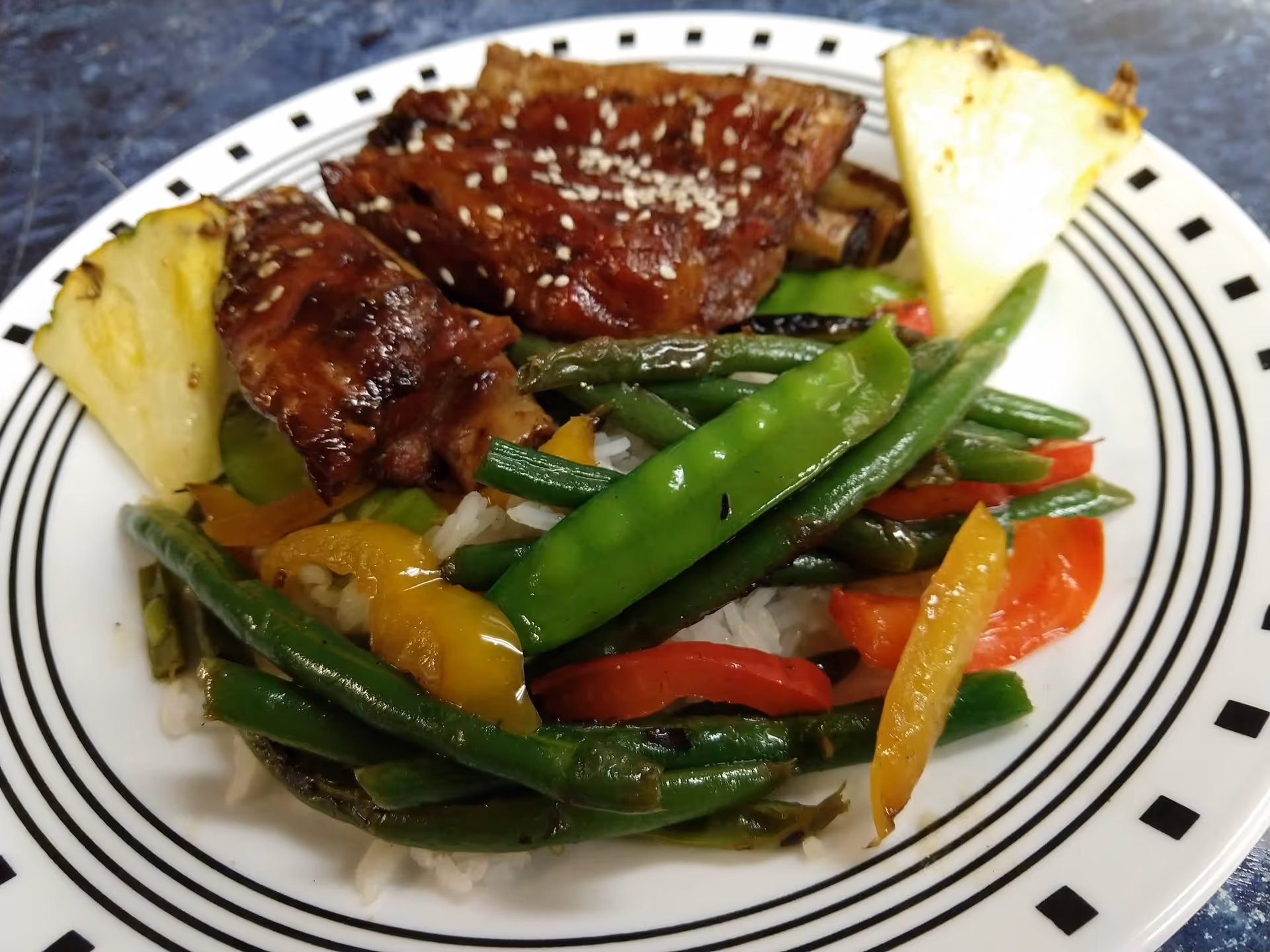 A plate of food featuring grilled ribs sprinkled with sesame seeds, served with sautéed green beans, red and yellow bell peppers, white rice, and two pineapple wedges on the side.