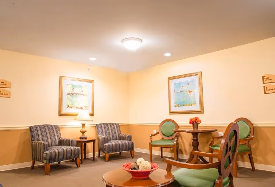 A cozy sitting area in a senior living facility with two striped armchairs separated by a small wooden table with a lamp, and a round wooden table with three green cushioned chairs. The walls are painted beige with two framed paintings hanging above the chairs. The room is softly lit with ceiling lights.