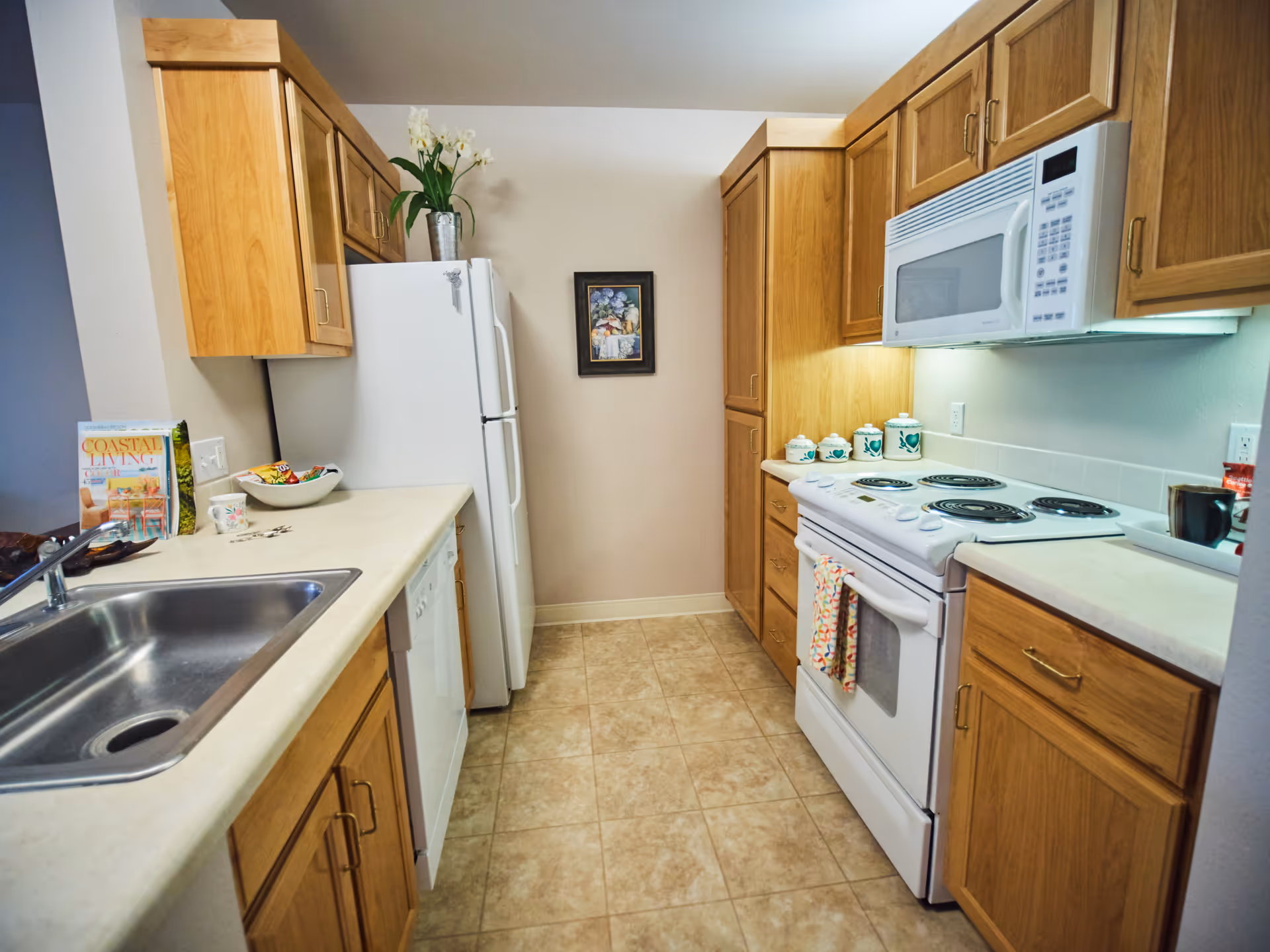 A compact kitchen with light wood cabinets, a white refrigerator, white electric stove with oven, white microwave, and a stainless steel sink. The countertops are beige, and there are decorative items including a bowl of snacks, a magazine, a flower vase on top of the refrigerator, and ceramic containers on the counter. The floor is tiled with beige tiles, and a framed picture hangs on the wall at the end of the kitchen.