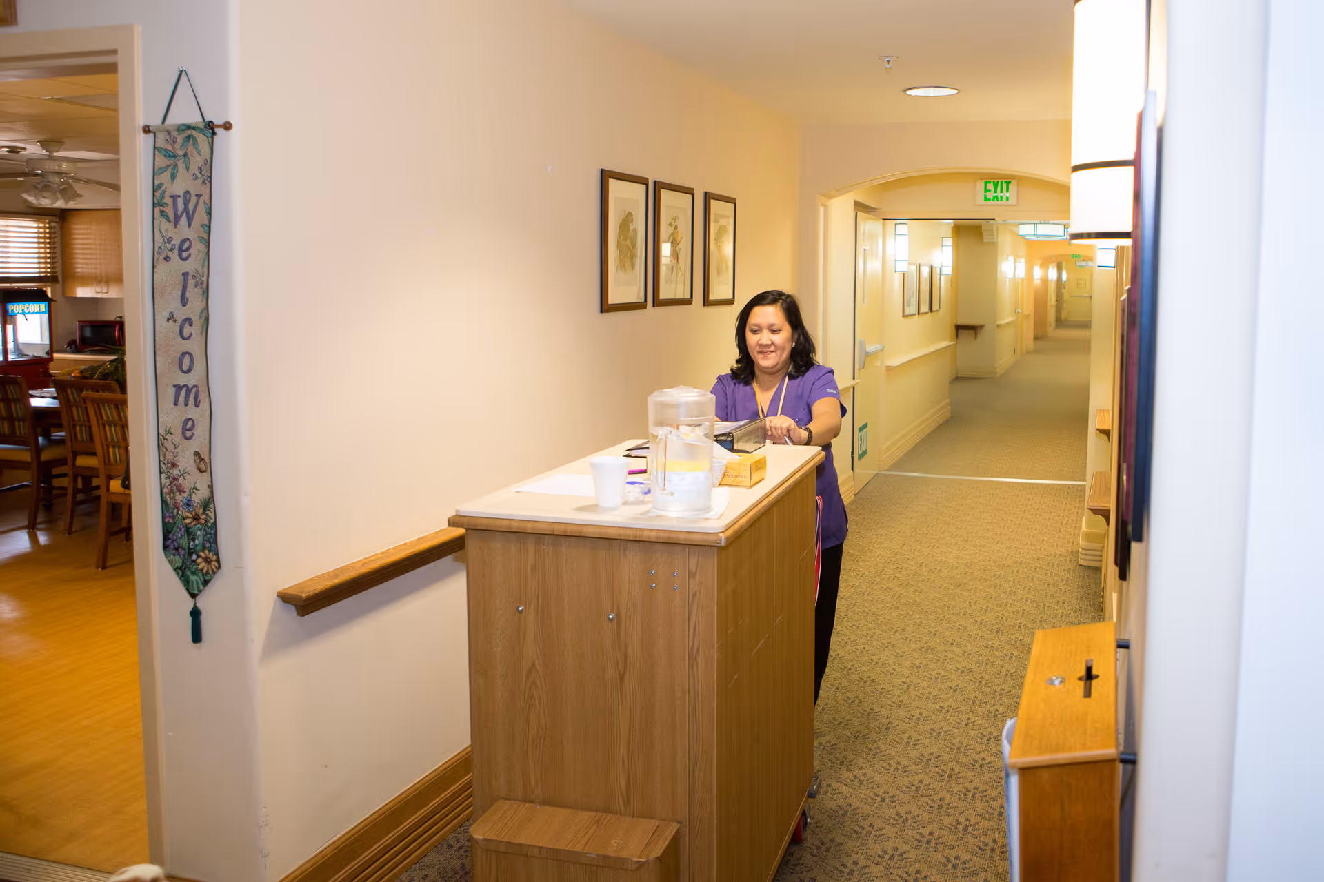 A woman in a purple uniform stands behind a wooden cart in a hallway of a senior living facility. The hallway is carpeted and has framed pictures on the walls. To the left, there is an open doorway leading to a dining area with wooden chairs and tables. A vertical 'Welcome' banner hangs on the wall near the doorway.