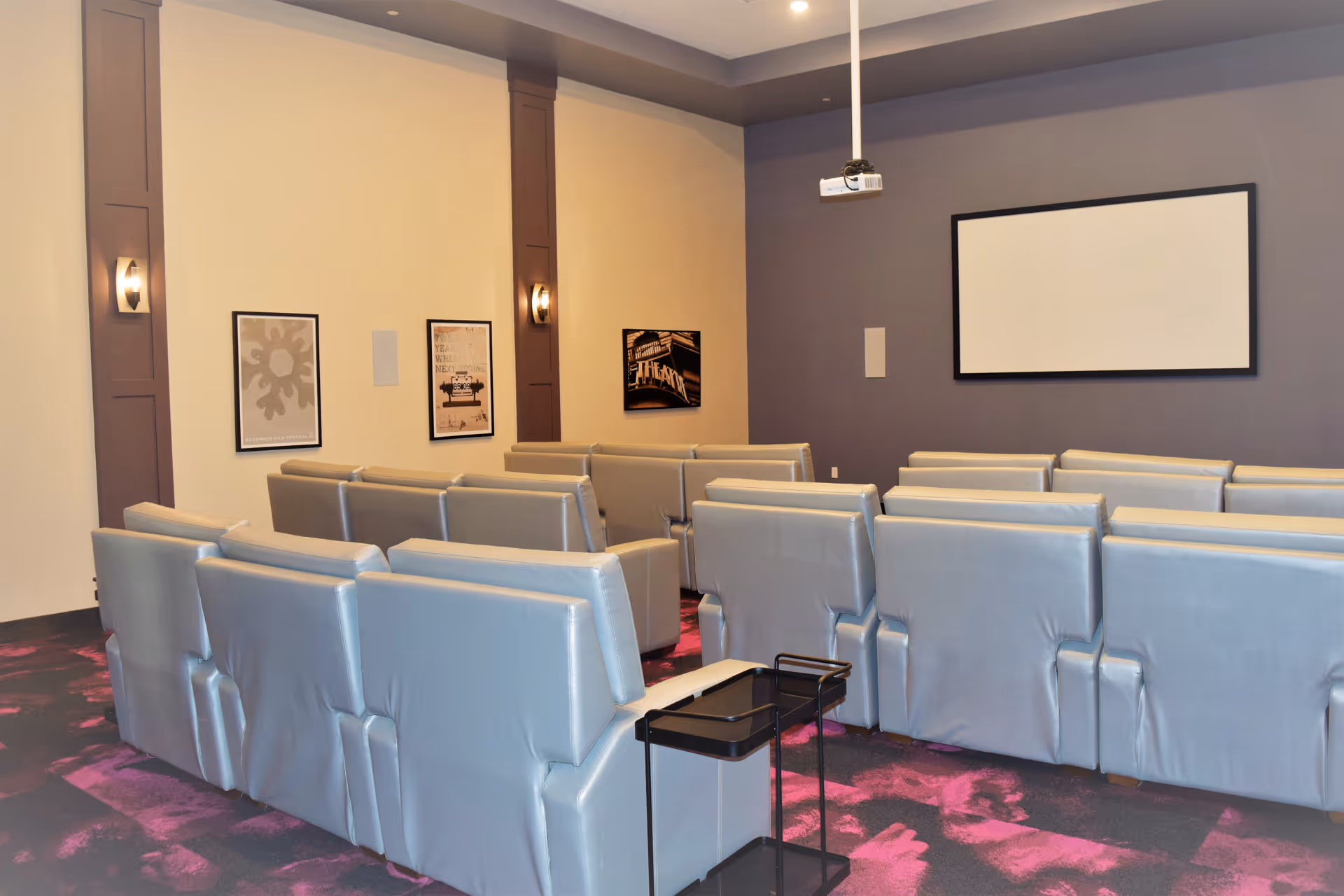 A small screening room with rows of gray reclining chairs facing a blank projection screen and ceiling-mounted projector.