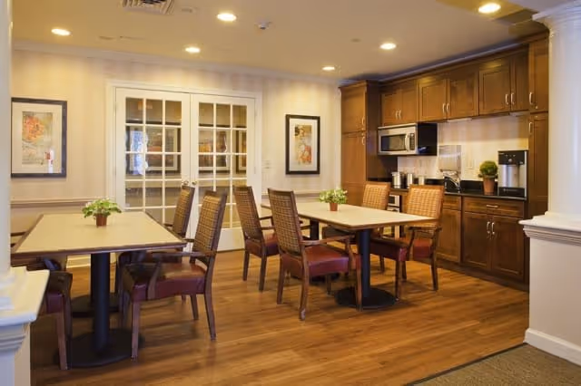 A cozy dining area with wooden floors, two rectangular tables each surrounded by four wooden chairs with red cushions. The room features a kitchenette with dark wood cabinets, a microwave, coffee maker, and a small plant on the counter. Two framed pictures hang on the wall beside white double doors with glass panes. The ceiling has recessed lighting.