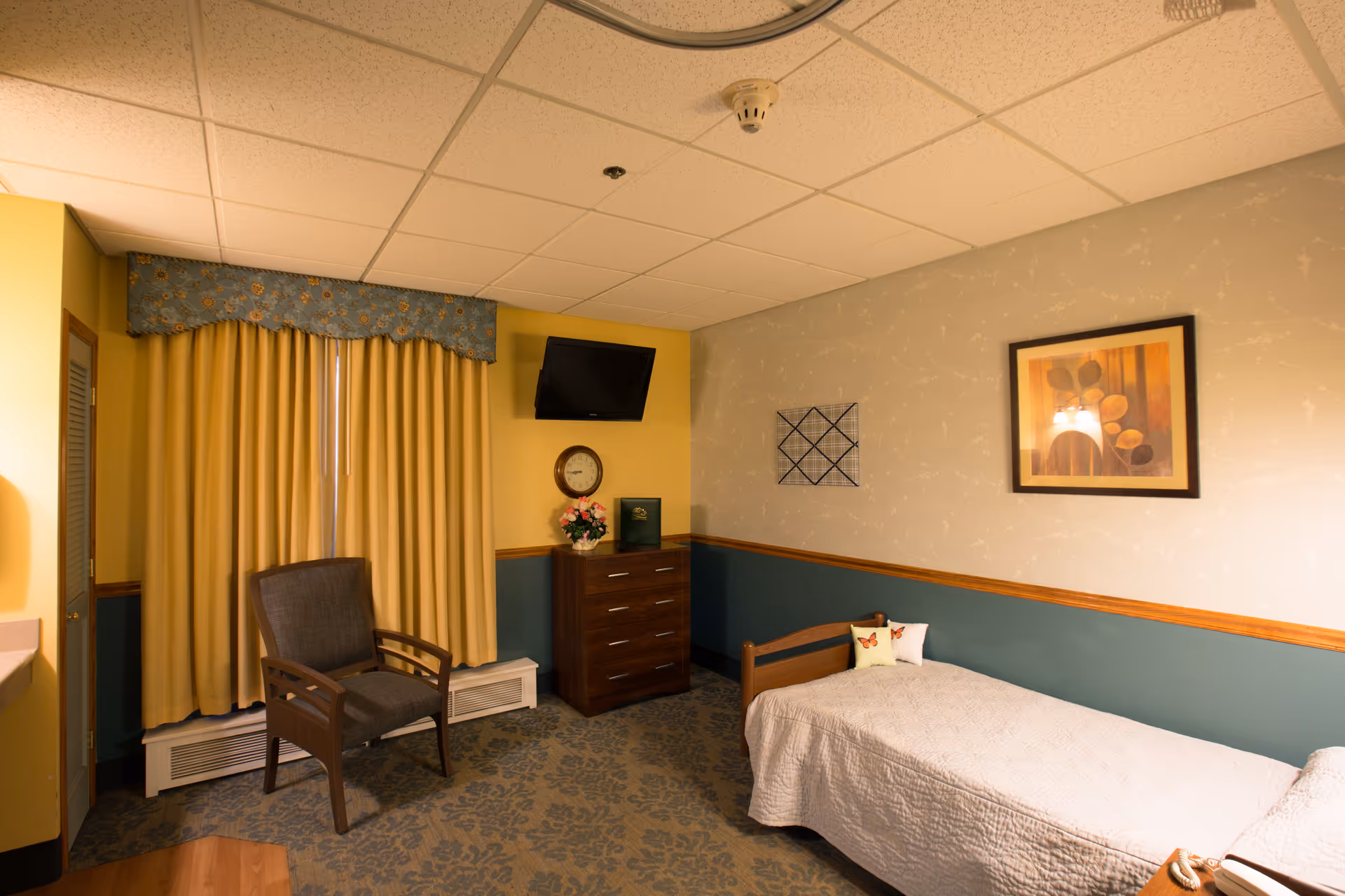 A cozy bedroom in a senior living facility with a single bed covered in a light quilt, a wooden chair, a chest of drawers with a clock and flower vase on top, a wall-mounted TV, and framed artwork on the walls. The room has yellow and blue walls with a patterned carpet and yellow curtains covering the window.