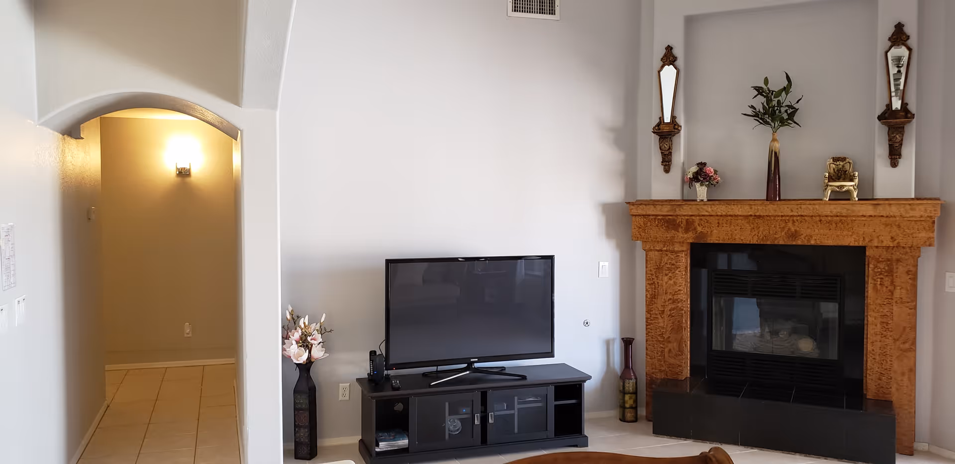 A living room featuring a flat-screen TV on a stand next to a decorative wood fireplace mantle and an arched hallway entrance.