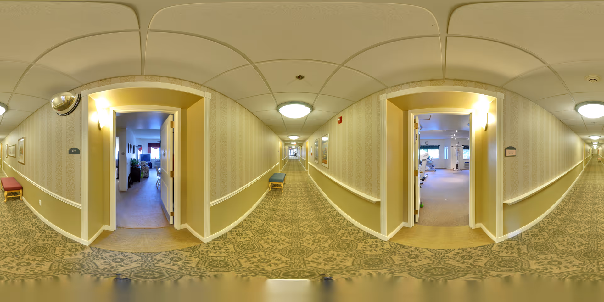 A long, carpeted hallway in a senior living facility with patterned wallpaper and ceiling lights. Two open doorways reveal interior rooms furnished with chairs, tables, and plants. Benches are placed along the hallway walls.