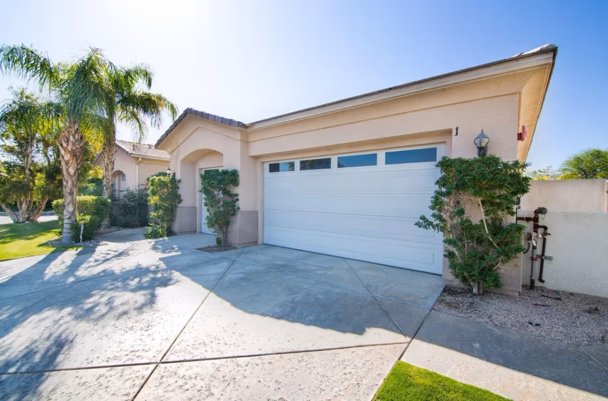 Exterior view of a single-story building with a white garage door, beige walls, and a tiled roof. There are palm trees and green shrubs around the driveway, and the sky is clear and blue.