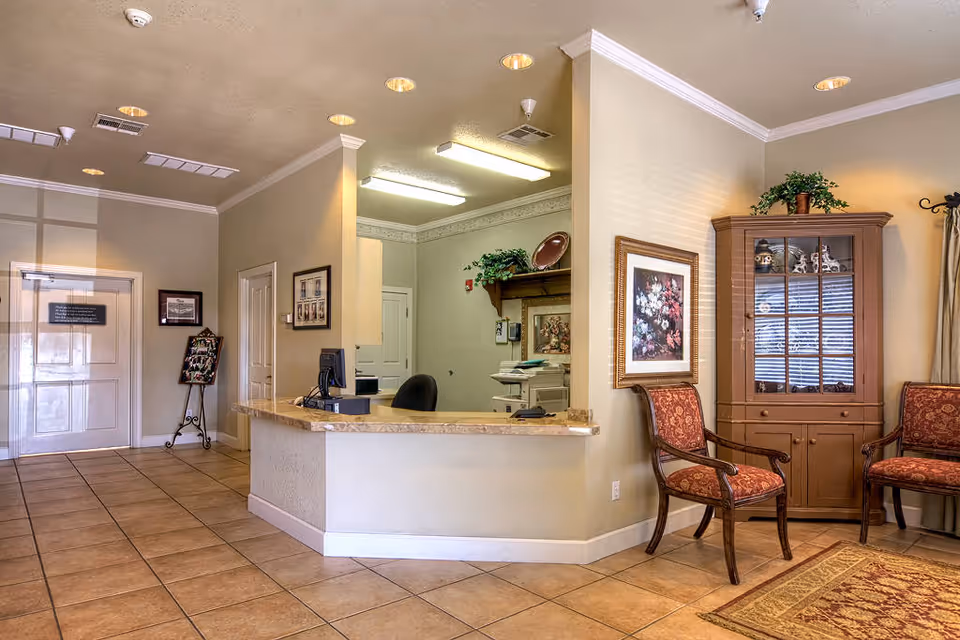 Reception area of Desert Hills Memory Care Center featuring a beige tiled floor, a marble countertop reception desk with a computer and office chair behind it, beige walls with framed artwork, a wooden cabinet with decorative items, two upholstered chairs with red patterned fabric, and ceiling lights.
