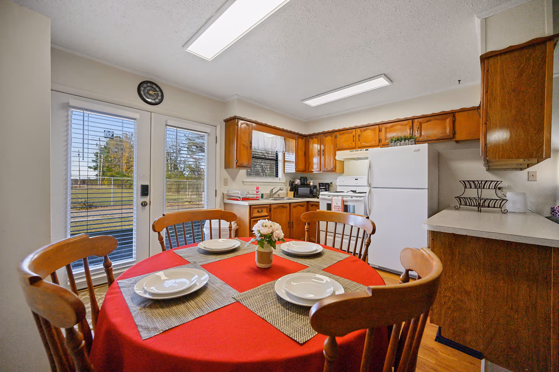 A cozy kitchen and dining area with wooden cabinets, a white refrigerator, stove, and sink. A round dining table covered with a red tablecloth is set with four plates and placemats, and a small flower vase is in the center. French doors with blinds lead outside, and a clock is mounted above the doors.