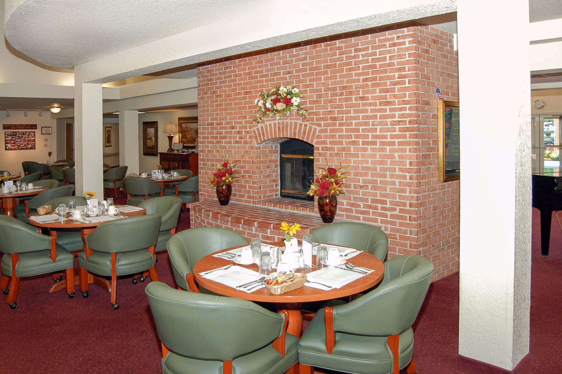 Dining area with round wooden tables set for meals, surrounded by green cushioned chairs. A large brick fireplace decorated with flower vases and a floral arrangement is centrally located. The room has a red carpet and white walls with columns.