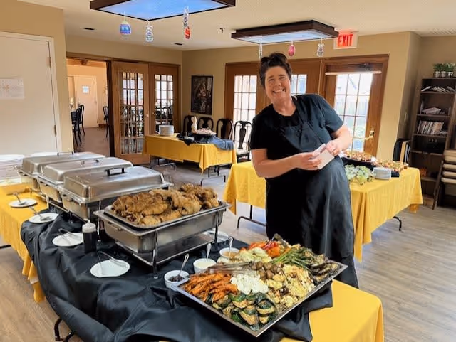 A woman dressed in black stands smiling behind a buffet table filled with trays of food including roasted vegetables and fried items in a dining area with yellow tablecloths and wooden chairs.