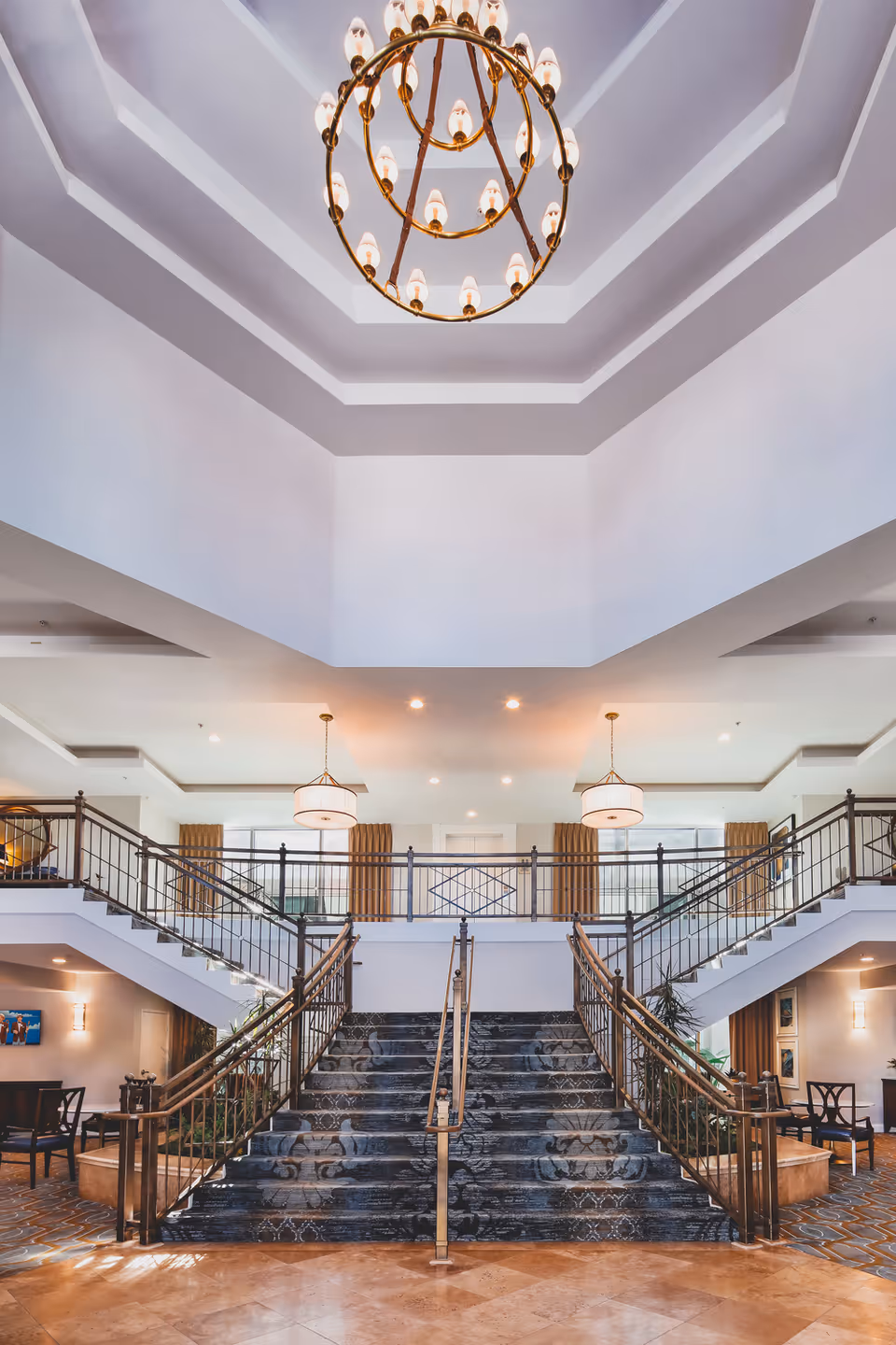 Spacious two-story interior lobby with a central double staircase, hanging chandelier, and seating areas.
