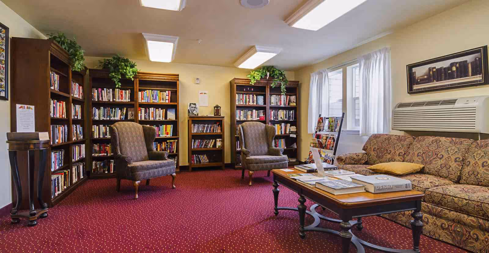A cozy library room with red carpet, two upholstered armchairs, a patterned sofa, and a wooden coffee table with books on it. The walls are light yellow, and there are several wooden bookshelves filled with books. A window with white curtains and an air conditioning unit is on the right wall. Green plants are placed on top of the bookshelves.