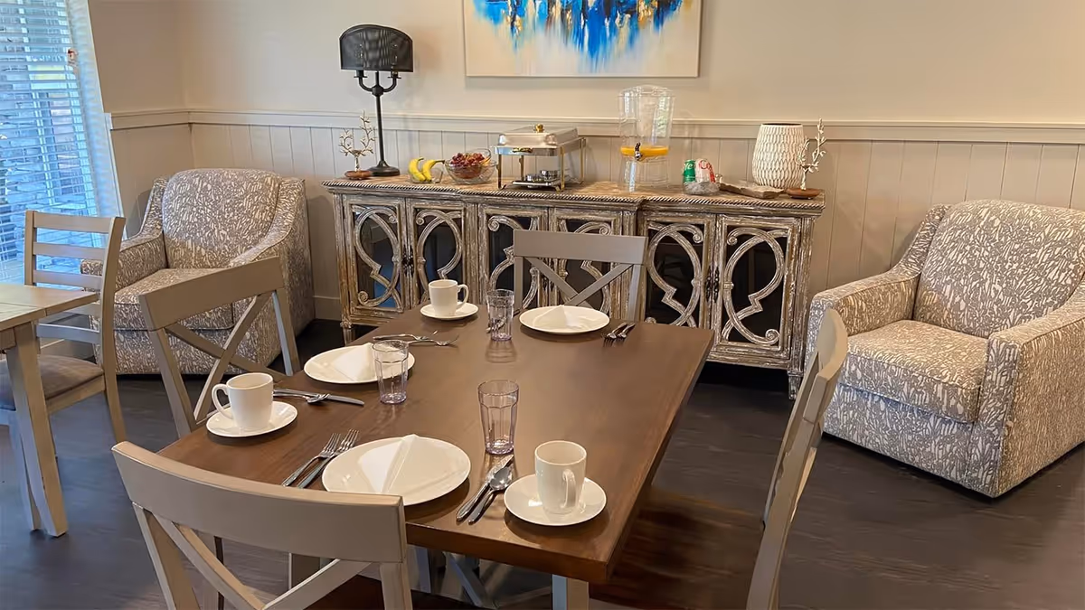 A dining area with a wooden table set for four with white plates, cups, and glasses. Surrounding the table are four chairs with light-colored frames. In the background, there are two patterned armchairs and a decorative sideboard with various items including a lamp, fruit bowl, and beverage dispenser. A colorful abstract painting hangs on the wall above the sideboard.