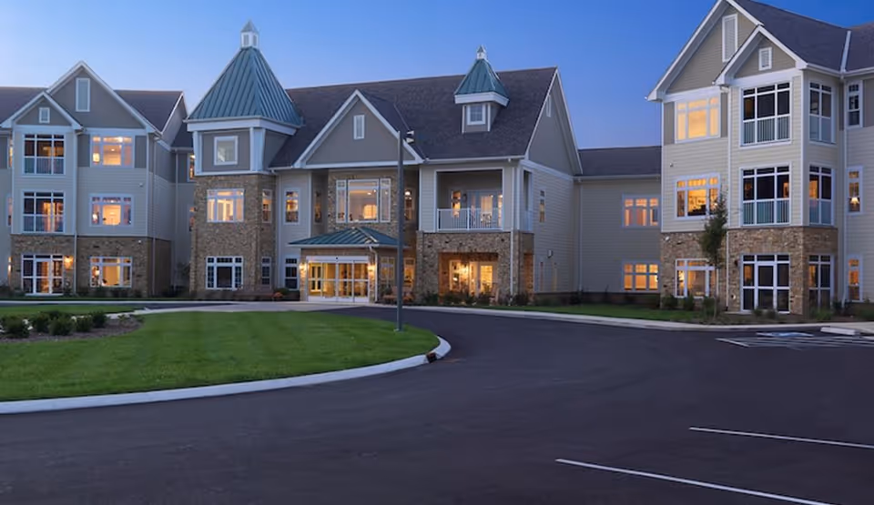 Exterior view of a large, multi-story senior living facility building at dusk with lights glowing warmly from the windows. The building features a combination of stone and siding with peaked roofs and a circular driveway in front surrounded by well-maintained grass and landscaping.