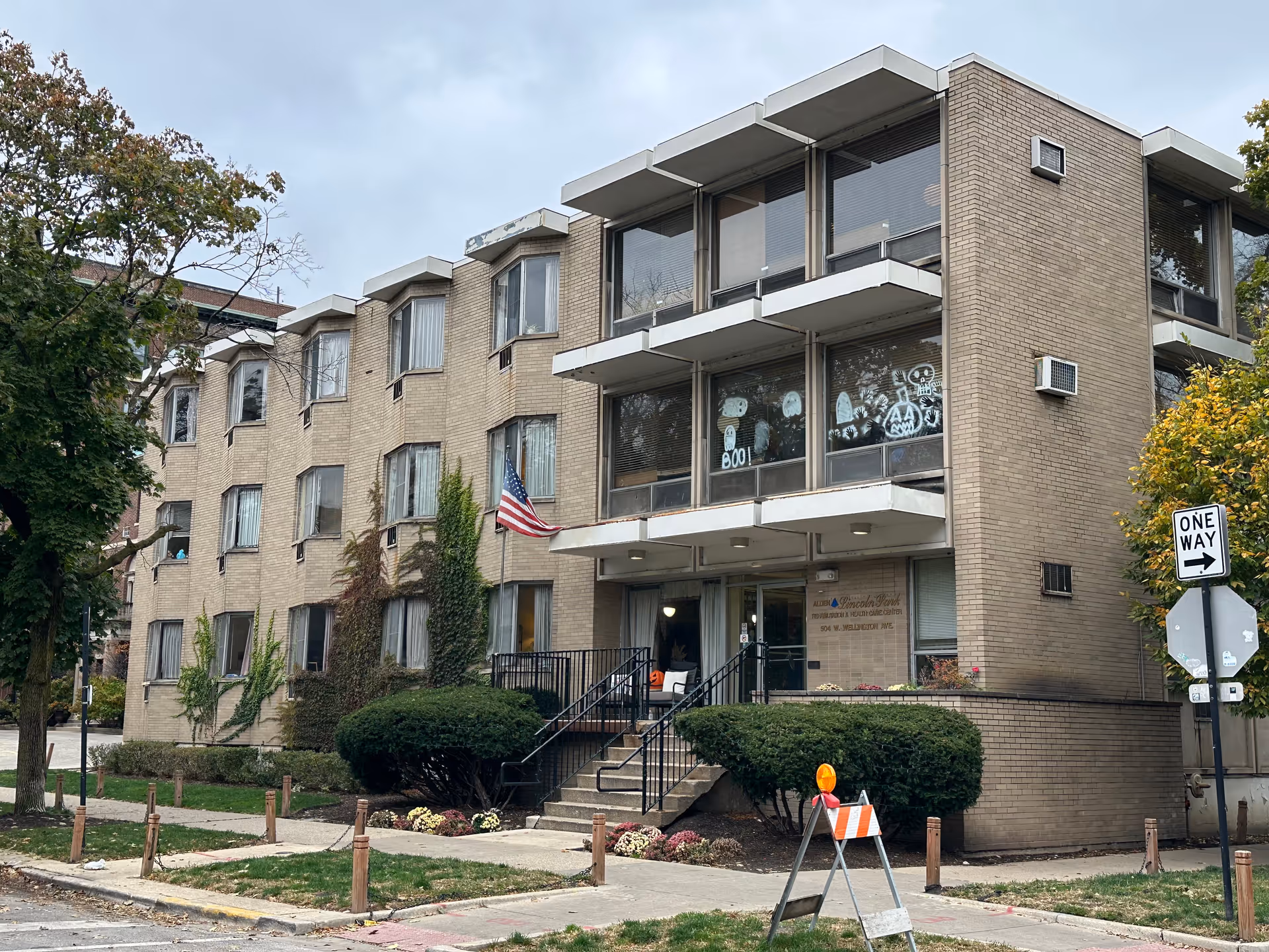 Exterior view of a three-story brick building with multiple windows, some decorated with Halloween-themed drawings. There is a small staircase leading to the main entrance, flanked by bushes and an American flag mounted on the wall. A one-way street sign is visible on the right side of the image, along with trees and a sidewalk.