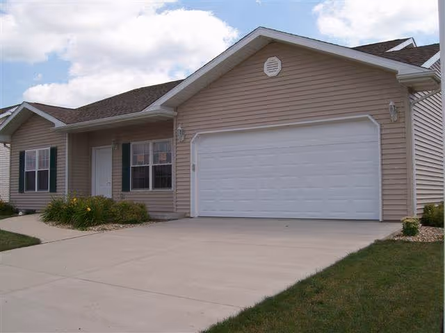 Exterior view of a single-story residential building with beige siding, a white garage door, a white front door, and windows with dark green shutters. There is a concrete driveway and a small lawn with some landscaping.
