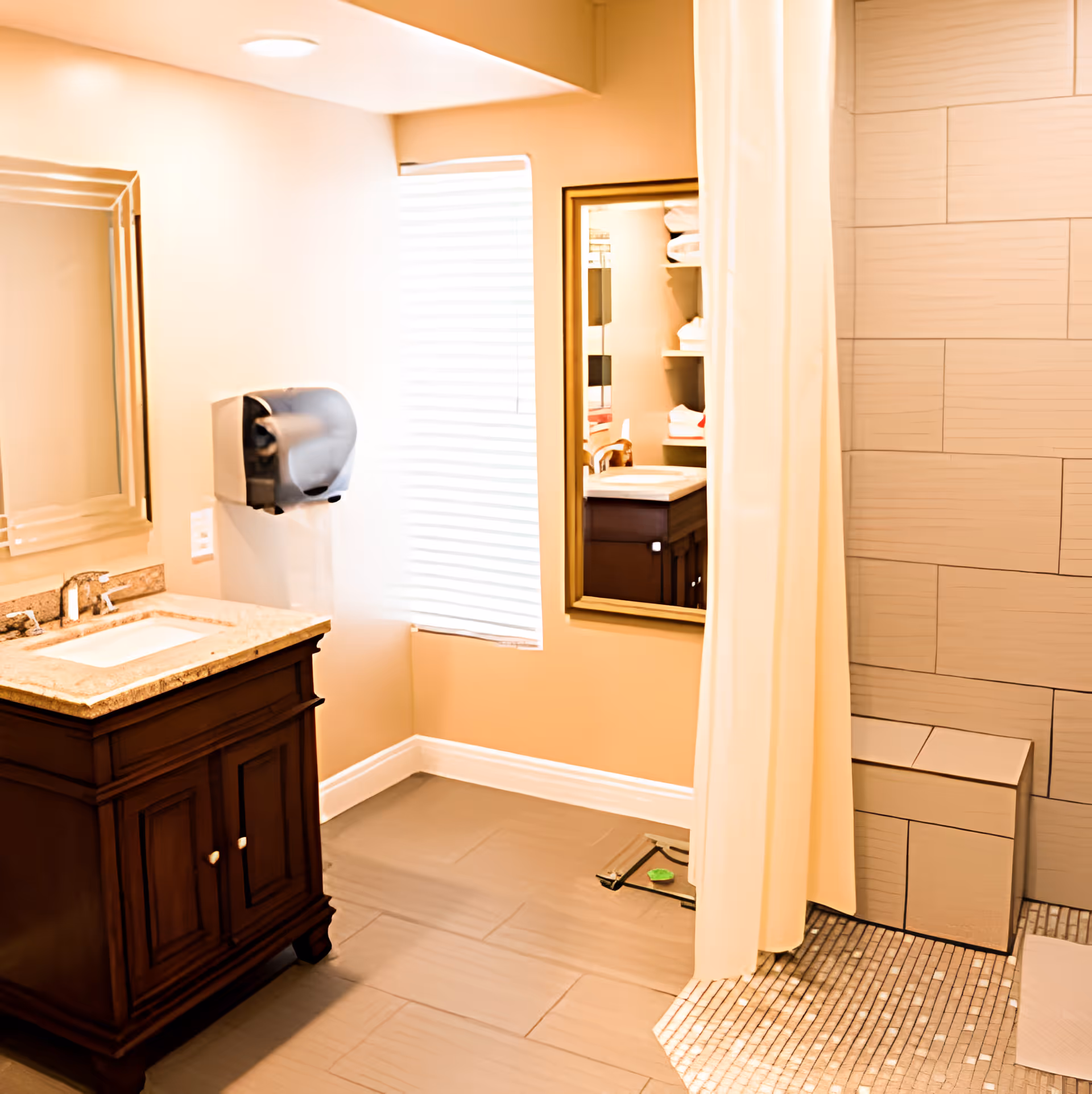 Bright bathroom with a wooden vanity featuring a granite countertop and sink, a large mirror above it, a paper towel dispenser on the wall, a window with blinds, a wall-mounted mirror reflecting shelves with towels, and a walk-in tiled shower area with a bench and a white shower curtain.