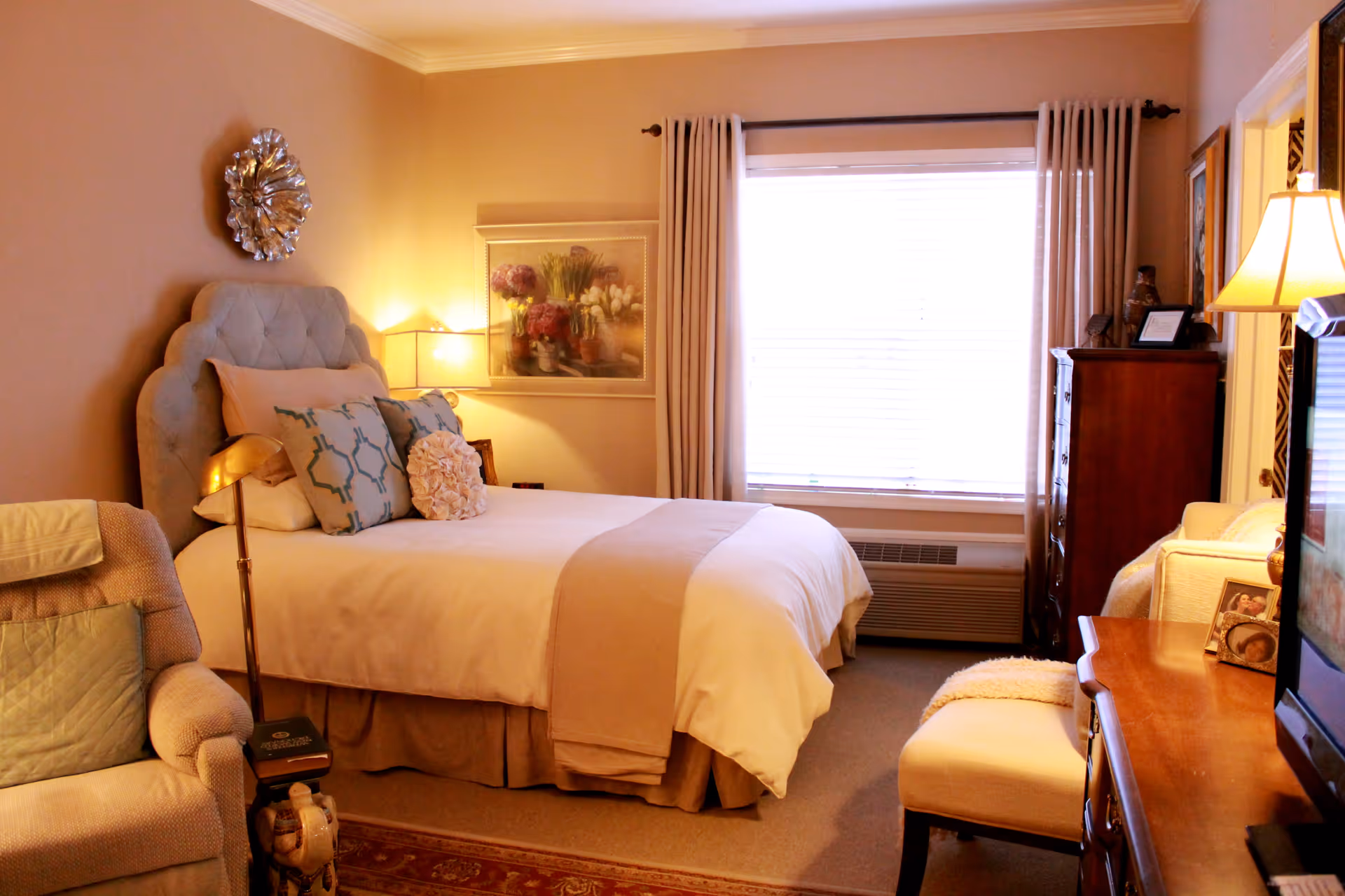 A cozy bedroom with a neatly made bed featuring multiple pillows and a beige throw blanket. There is a cushioned armchair with a pillow on the left side and a wooden dresser with a lamp and framed photos on the right. A large window with curtains lets in natural light, and a decorative wall piece and framed artwork are visible on the walls.