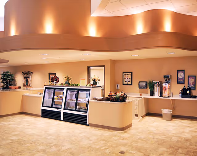 A warm-toned dining service area with a refrigerated display case, coffee station, and serving counters under curved ceiling lighting.