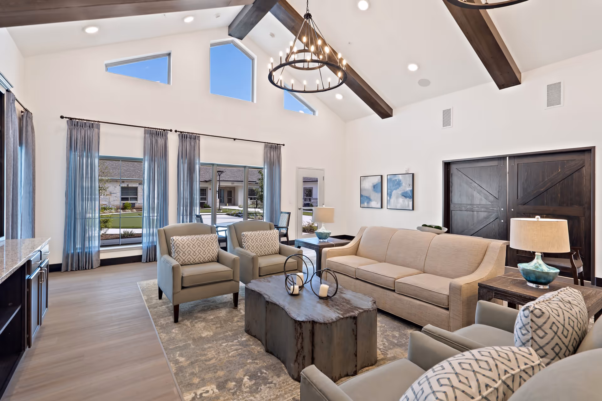 A bright and spacious living room with high vaulted ceilings featuring dark wooden beams and a circular chandelier. The room has large windows with blue curtains allowing natural light to fill the space. There is a beige sofa, two beige armchairs with patterned cushions, a rustic wooden coffee table with decorative candle holders, and side tables with lamps. The floor is light wood, and there are two abstract paintings on the wall next to dark wooden double doors. Outside the windows, a courtyard with greenery is visible.