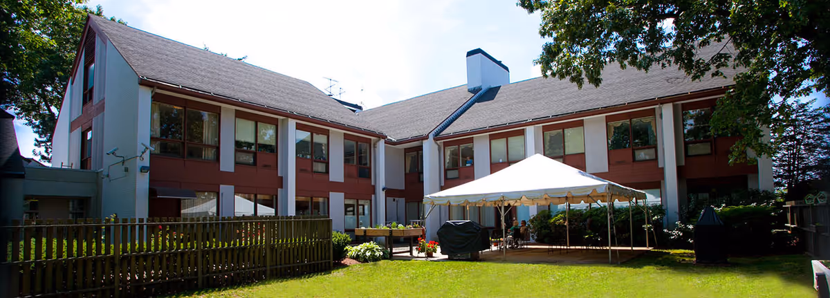 Exterior view of a two-story senior living facility building with large windows and a pitched roof. In front of the building, there is a white canopy tent set up on a grassy lawn, surrounded by some outdoor furniture and a wooden fence on the left side. Trees and shrubs are visible around the building.