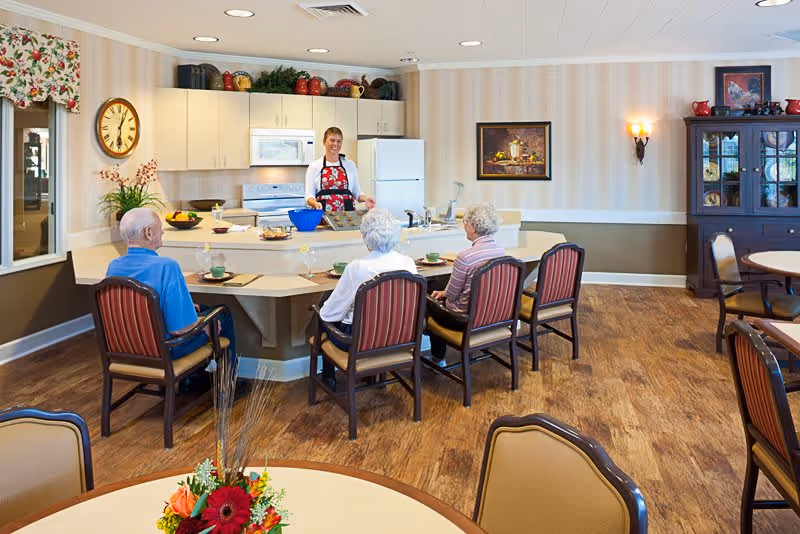 A kitchen and dining area in a senior living facility with a woman standing behind a kitchen counter preparing food. Four elderly people are seated on chairs at the counter, facing her. The room has wooden floors, beige walls with striped wallpaper, and various decorations including a clock, framed pictures, and a cabinet with dishes.