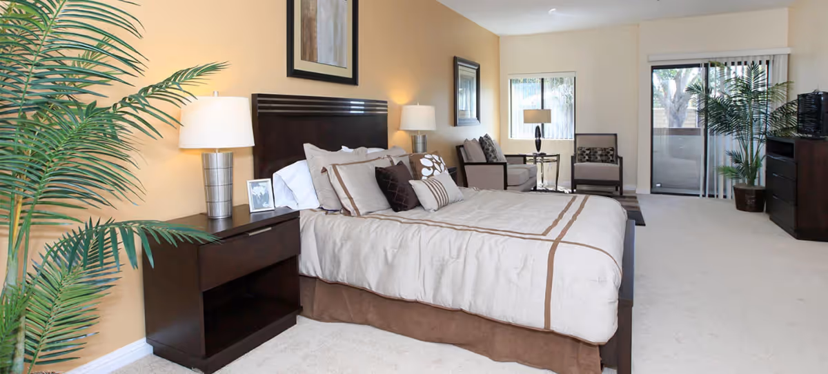 A spacious bedroom with a large bed featuring beige and brown bedding, two bedside tables with lamps, a framed picture on the wall, a sitting area with a sofa and chair, large windows, sliding glass door leading to a balcony, and potted plants.