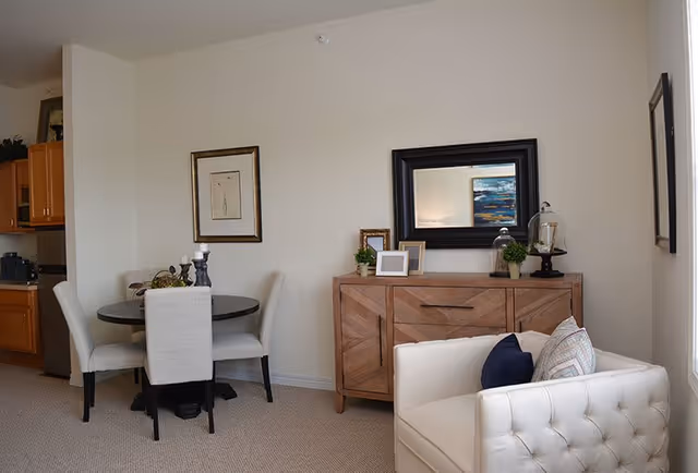 A cozy interior space featuring a small round dining table with four upholstered chairs, a wooden sideboard with decorative items including framed pictures, plants, and a mirror above it, and a white tufted armchair with cushions. The room has neutral walls and carpeted flooring, with a partial view of a kitchen area in the background.