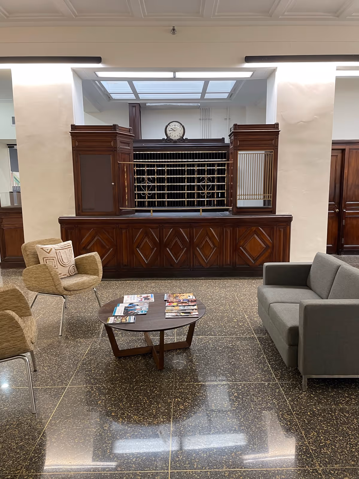 A waiting area with a gray sofa, two beige armchairs with patterned cushions, and a small wooden coffee table with magazines on it. Behind the seating area is a vintage wooden reception desk with metal mail slots and a clock on top. The floor is polished with a speckled pattern, and the walls are light-colored.